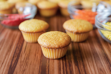 Three mini cupcakes with glass bowls of red, orange and yellow sprinkles on a wood surface.