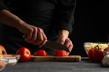 the chef cooks, slices tomatoes for cooking. on a black background. recipe book and tasty wholesome food. cooking specials