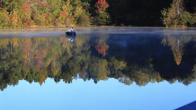 A lone fisherman drifts through the reflections of autumn trees on a chilly morning at the lake.