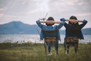 Rear view of two happy young gir sitting relaxed put hands behind her head a park, on amidst nature and water in a park , travel concept, Cheerful Concept Friendship, Camping and travel concept.