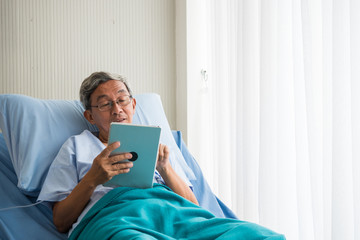 Happy elderly patient sitting on bed and making video call with tablet at hospital.