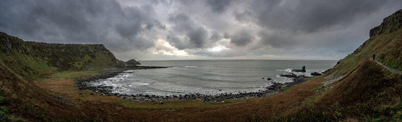 Fototapeta na wymiar Panoramic view of the pathways and the bay at the coast called Giant's Causeway, a landmark in Northern Ireland, with the cliffs that surround the place and dramatic cloudy sky.