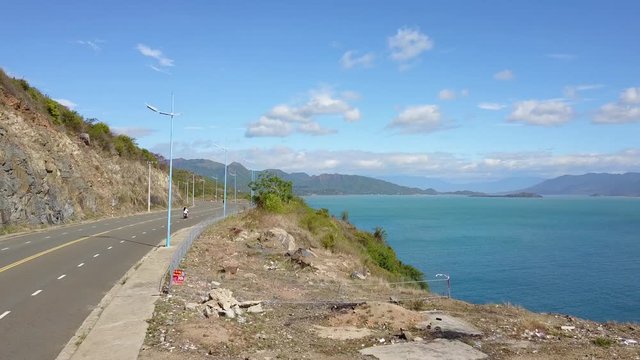 A flying camera takes a motorcyclist passing on a road near the sea with a view of the rocky shore.