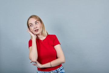 Portrait of young woman in red shirt eavesdrop with hearing gesture, looking aside isolated on grey...