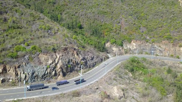 A flying camera takes a motorcyclist passing on a road near the sea with a view of the rocky shore.