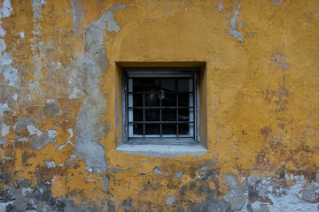 Fragment of an old plastered wall in yellow. In the middle of the wall there is a square window with bars. Background. Texture.