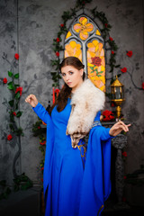 Young beautiful girl in a medieval blue dress with long sleeves and fur on her shoulders posing against the backdrop of a window, fireplace and flowers.