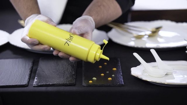 Camera shows man's hands decorating black rectangular plate by putting drops on it from yellow plastic bottle on table somewhere in restaurant preparing to catering.