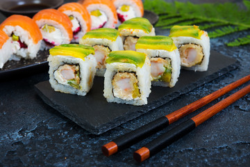 A set of sushi rolls with avocado and shrimp lies on a black charcoal Board. Traditional Japanese food with chopsticks