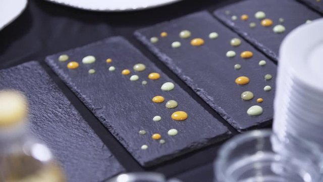 Camera shows man's hands making decoration of black rectangular plate by putting drops on it from red plastic bottle on table somewhere in restaurant preparing to catering.