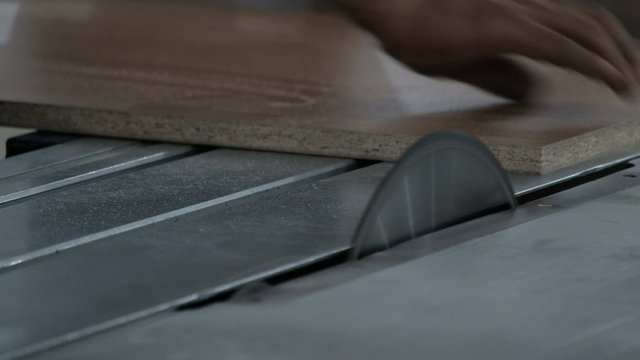 The worker saws off the edge of the chipboard on a machine with a circular saw. Close-up. Furniture manufacture. The video contains real production sound and noise.