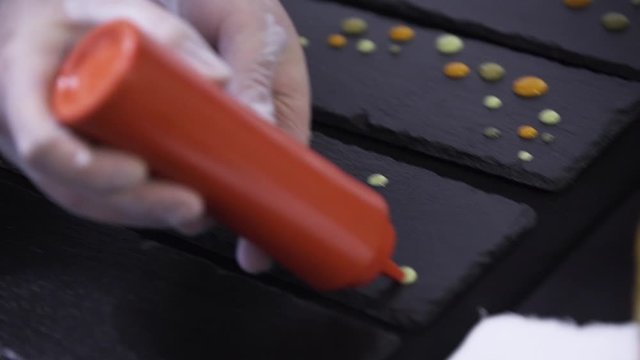 Camera shows man's hands making decoration of black rectangular plate by putting drops on it from red plastic bottle on table somewhere in restaurant preparing to catering.