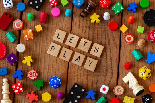 "Lets Play" spelled out in wooden letter tiles. Surrounded by dice, cards, and other game pieces on a wooden background