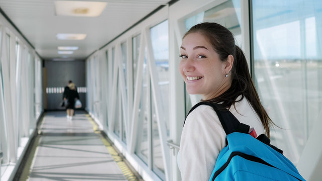 Happy young woman with backpack is going on telescopic gangway to the airplane. She turns around, looks at the camera and smiles.