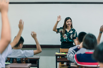 An smiling Asian female high school teacher teaches the white uniform students in the classroom by asking questions and then the students raise their hands for answers.