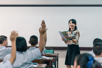 An smiling Asian female high school teacher teaches the white uniform students in the classroom by asking questions and then the students raise their hands for answers.
