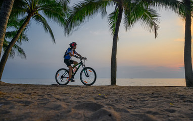 Fototapeta premium woman riding bicycle along the sea beach under coconut trees at sunset, relaxation at the end of the day.