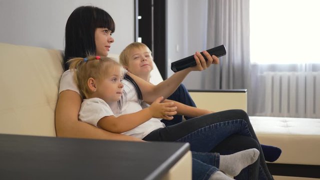 Happy family mom and two children are sitting on the couch in front of the TV. Smart TV switching remote control in the hands of a girl. Family watching television. Modern technology concept