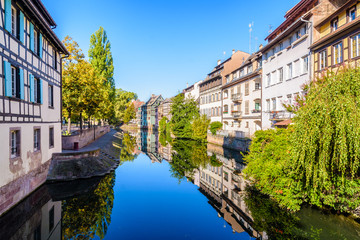 The river Ill canal bathes the Petite France quarter in Strasbourg, France, lined with half-timbered houses reflecting in the still waters under a bright sunshine.
