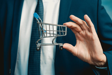 Male hands of a businessman in a blue suit with a tie hold a mini shopping trolley. 