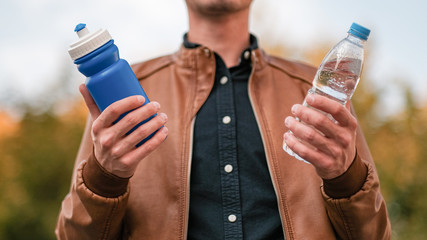 A young guy in yellow sunglasses holds a plastic bottle in one hand and a refillable bottle in the...