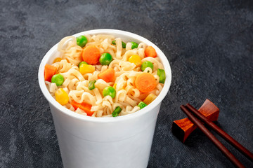 Ramen cup close-up, instant soba noodles in a plastic cup with vegetables, on a black background with chopsticks