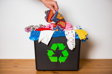 Hand putting used sock in bin with recycling sign. Person in the house separating waste. Heap of colourful clothes in black box. Textile utilisation concept 
