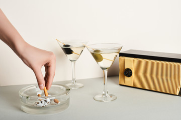Cropped view of woman putting cigarette to ashtray beside cocktails and vintage radio on white...