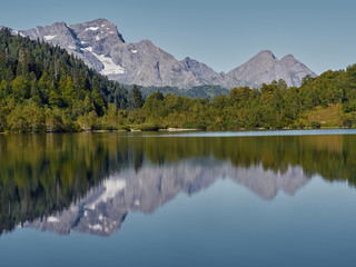 Panoramic view of Kardyvach lake in sunny morning among Caucasus mountains; mirror water surface reflecting autumn highland landscape, rocky peaks and overgrown steep slopes of Agepsta mountain ridges