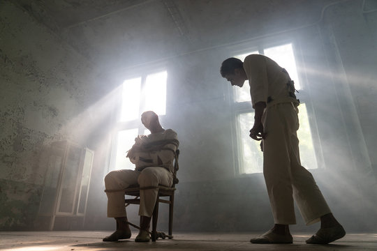 A crazy man in a straitjacket is tied to a chair in an abandoned old clinic and the other insane man coming closer with interest