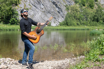 young hipster bearded guy, man in cap plays guitar with lowered strings against backdrop of bright summer landscape