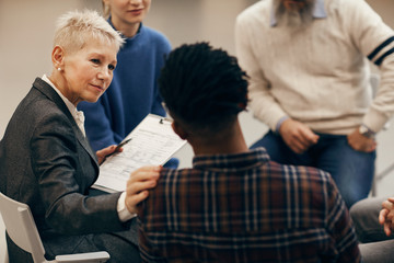 Mature woman with short blond hair listening to African young man and supporting him during therapy lesson with class