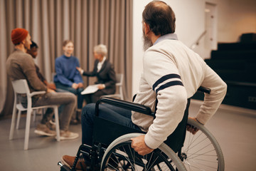 Obraz premium Rear view of senior man sitting in wheelchair and looking at people sitting and talking during therapy class