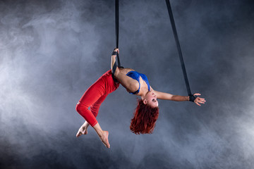 Female athletic, sexy and flexible aerial circus artist with redhead on aerial straps on black background
