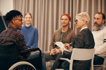 Young African man in wheelchair talking to mature woman while she making notes during therapy lesson