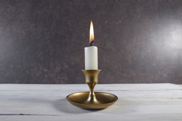 Burning candle in vintage metal candlestick on white wooden table against dark stone background..