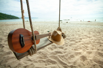 hat and ukulele placed on a swing on a white beach, blue sea, surrounded by blue sky with clouds, holiday travel concept