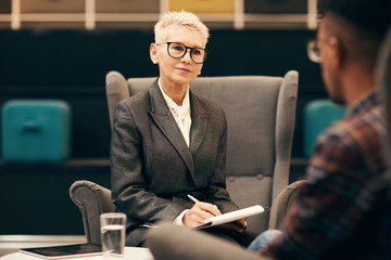 Mature businesswoman with short blond hair and in eyeglasses sitting and making notes while interviewing the young man during business meeting