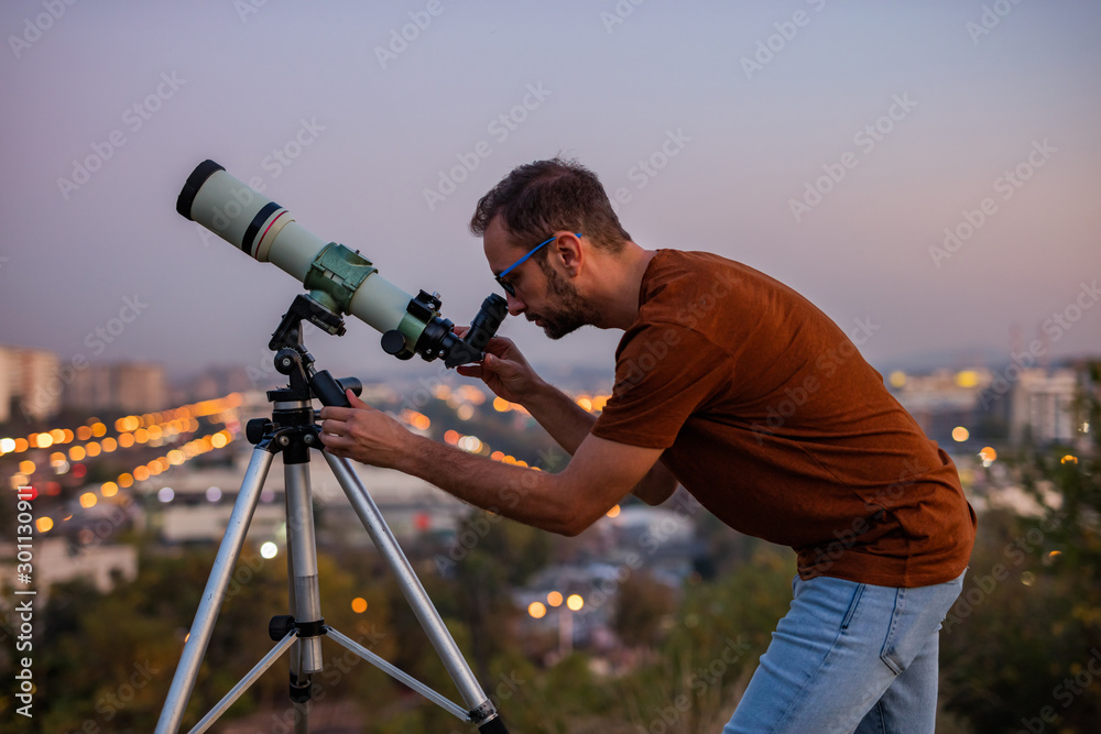 Wall mural astronomer with a telescope watching at the stars and moon with blurred city lights in the backgroun