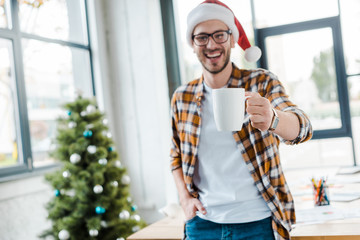 selective focus of happy bearded man in santa hat holding cup near christmas tree in office