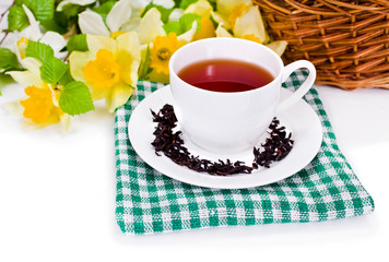 Black tea in a cup on green textile napkin with yellow daffodil flowers and green birch tree branches, wicker basket on a white background. Spring still life.