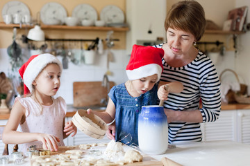 Family is cooking Christmas cookies together in cozy kitchen at home. Surprised kids and woman make holiday meal. Daughters help mother to bake gingerbreads. Candid moment. Children chef concept.