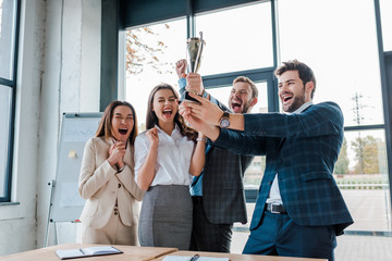 happy businessmen and multicultural businesswomen holding trophy in office - obrazy, fototapety, plakaty