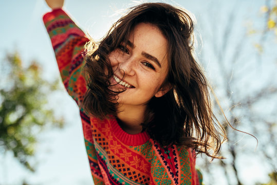 Bottom view close-up portrait of happy young woman smiling broadly with windy hair and freckles has joyful expression, wearing colorful knitted sweater with arms wide open, posing on nature sunlight