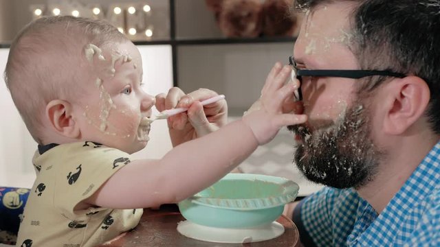 Father feeds baby from spoon. Dad and child have very dirty faces with porridge, baby behaves very messy, often puts his hand on plate and on face of parent. Close-up