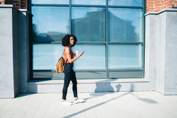 Fototapeta na wymiar Millennial hipster girl dressed in casual wear walking around city street with takeaway cup and smartphone gadget in hands,dark skinned female student strolling on urban setting with modern technology