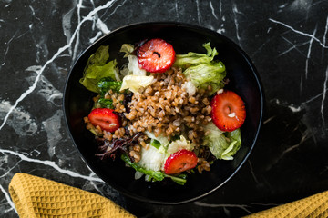 Buckwheat Salad with Strawberry Slices and Green Leaves in Black Bowl on Dark Granite Surface.