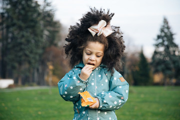 Little lovely girl on a walk eats cookies, a child eats healthy food, a happy kid walks on an autumn day, afro haircut