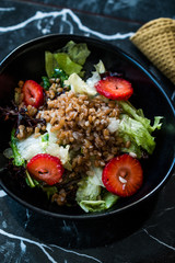 Buckwheat Salad with Strawberry Slices and Green Leaves in Black Bowl on Dark Granite Surface.