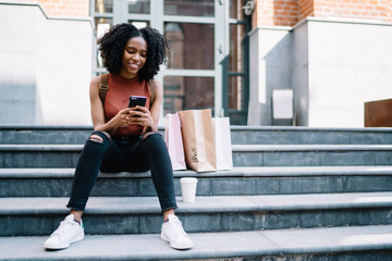 Obraz premium Cheerful hipster girl dressed in casual wear taking rest on stairs while checking email on smartphone device connected to 4g wireless for browsing websites, happy female buyer writing review